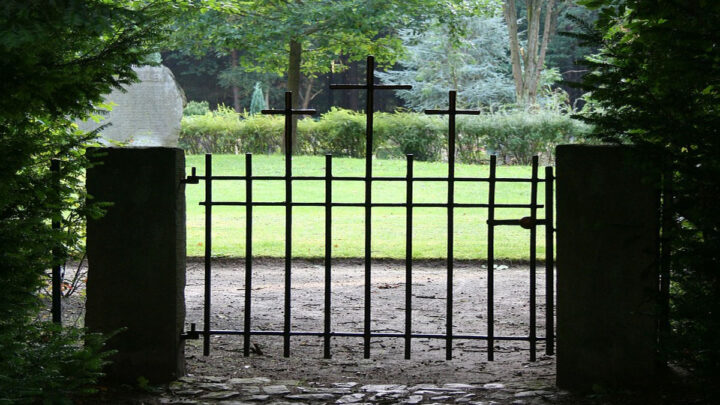 gate with 3 crosses and 2 brick columns on each side opening into a grass area surrounded by manicured bushes.